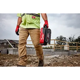 Construction worker carrying a Milwaukee toolbox, wearing safety gear, on a rocky construction site with buildings in the background.