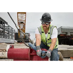 A construction worker wearing a hard hat, neon safety vest, and Full Frame Safety Glasses with Removable Side Shields – Silver Mirrored Anti-Scratch Lenses is kneeling while operating industrial equipment at a worksite featuring scaffolding and a crane.