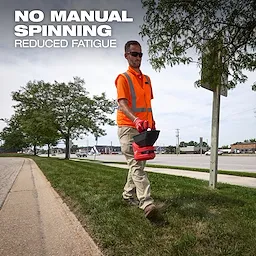 Worker walking along a grassy roadside carrying an M12 Handheld Broadcast Spreader in one hand.