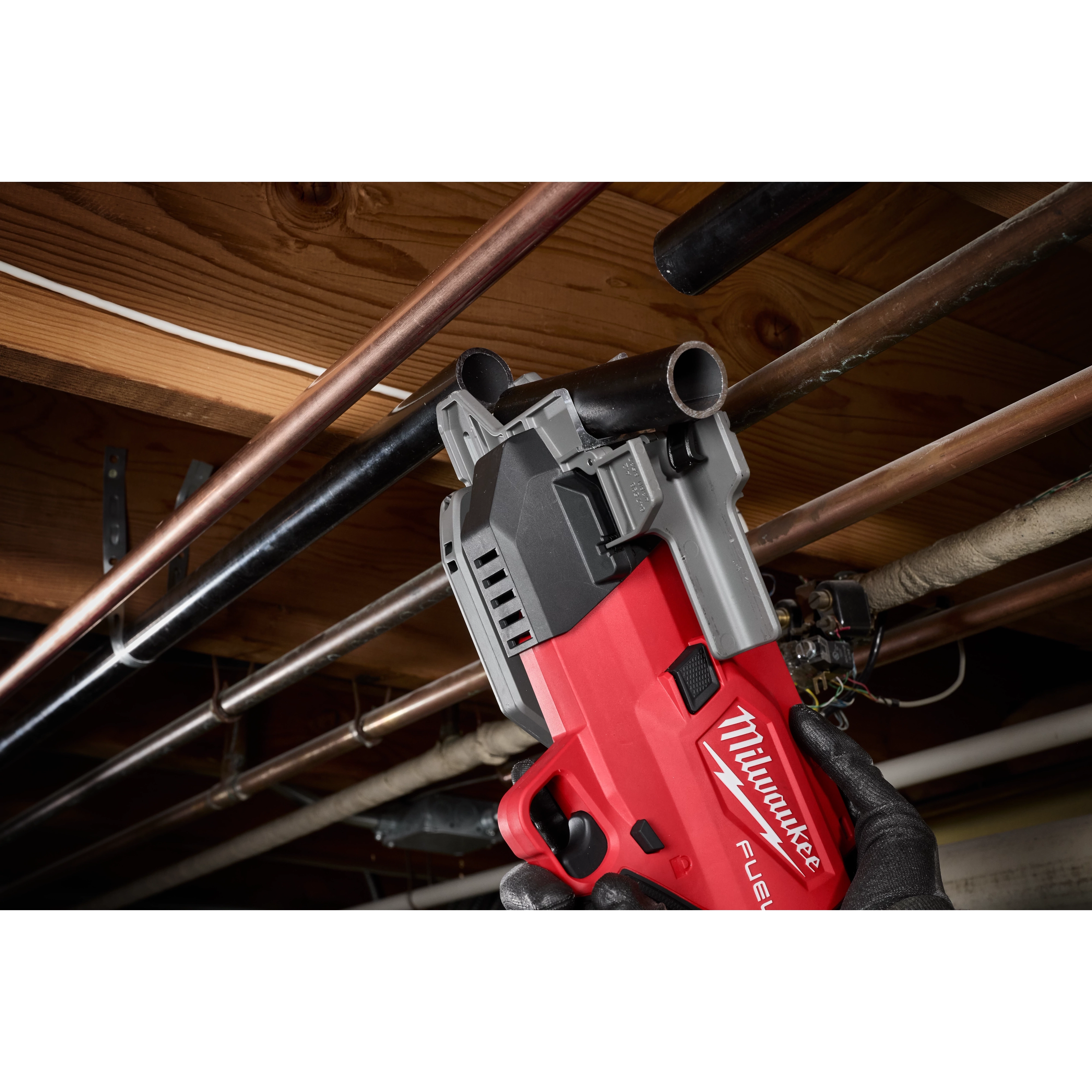 A person wearing gloves uses a red Milwaukee pipe cutter to cut through a black pipe in a basement with exposed wooden beams and various pipes.