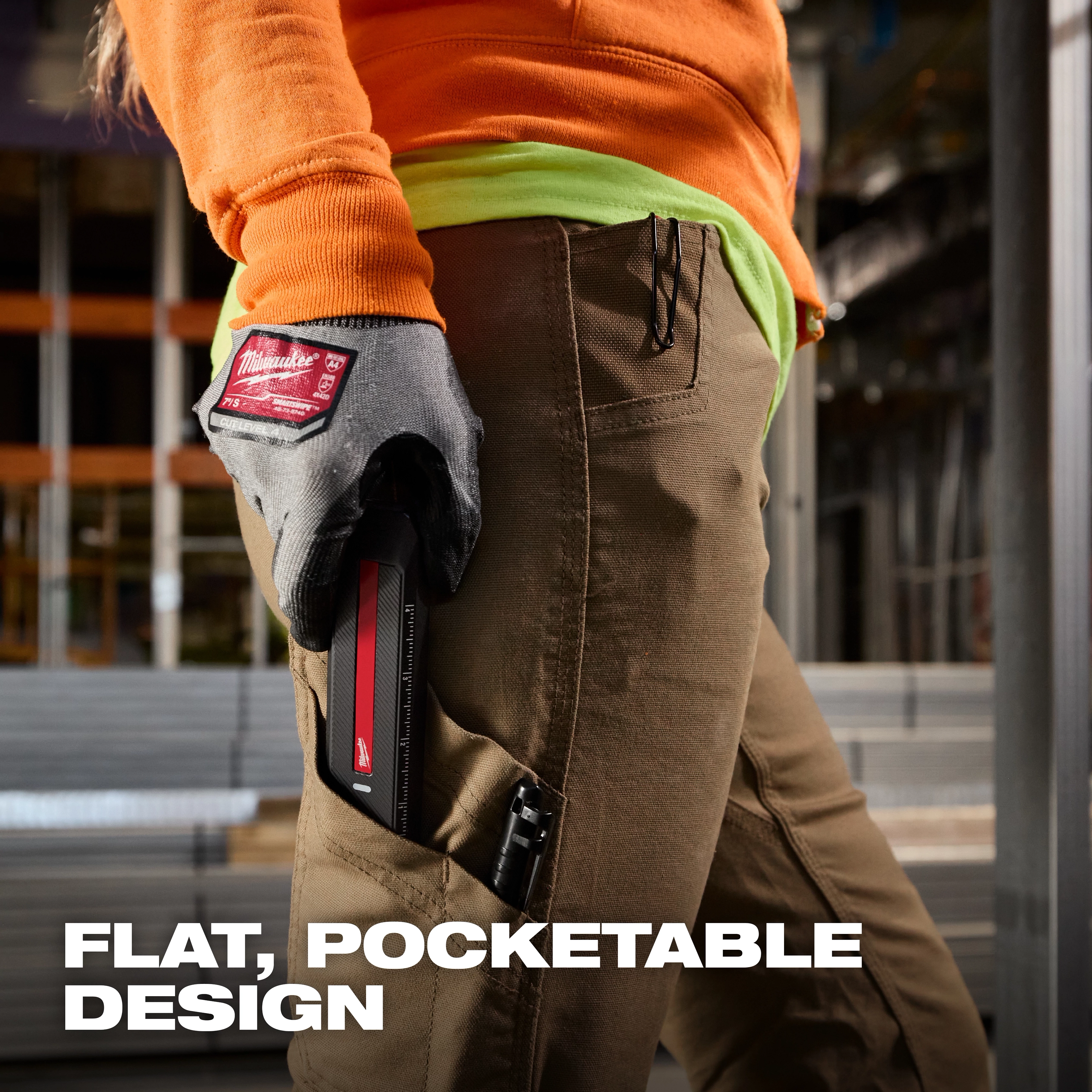 Gloved hand holding Milwaukee Rechargeable Flat Flashlight w/ Green Laser against work pants in an industrial setting.