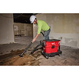 A person using the M18 FUEL™ NEXUS™ Dual Battery Wet/Dry Vacuum Motor Head with PACKOUT™ Compatibility & VACLINK™ to clean a construction site. The vacuum is red with a black attachment and is being used to remove debris from the floor.