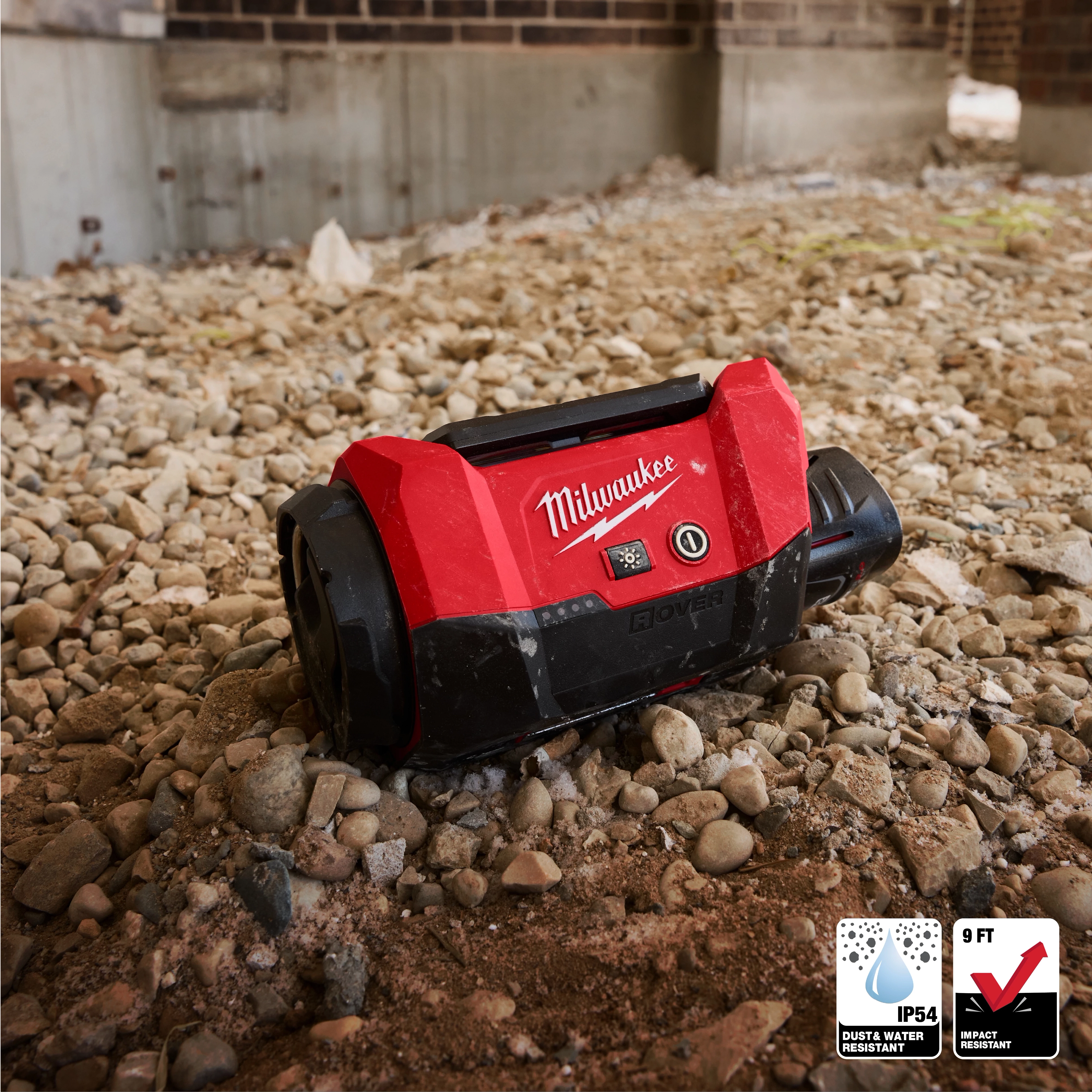 The Milwaukee M12™ ROVER™ Multi-Directional Flood Light with PACKOUT™ Compatibility sits on a rocky ground. It is red and black, designed for durability, water and dust resistance (IP64), and impact resistance up to nine feet. The background shows a construction site with exposed concrete and bricks.