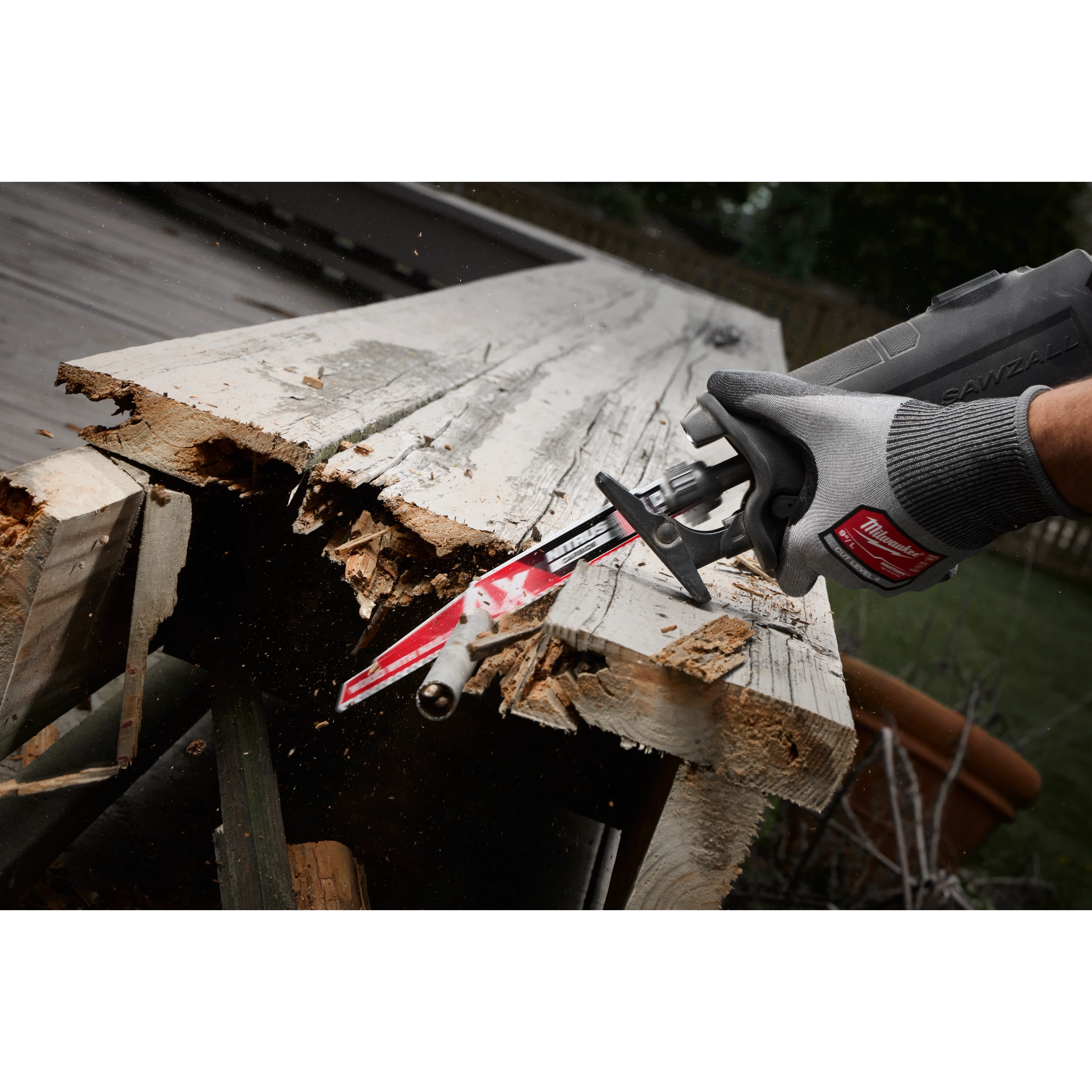 A person uses a 9" 5 TPI AX™ w/NITRUS™ Carbide Teeth SAWZALL® Blade for Wood with Nails to cut through a piece of weathered wood with embedded nails. The blade is attached to a power saw, demonstrating its capability in cutting through tough materials.