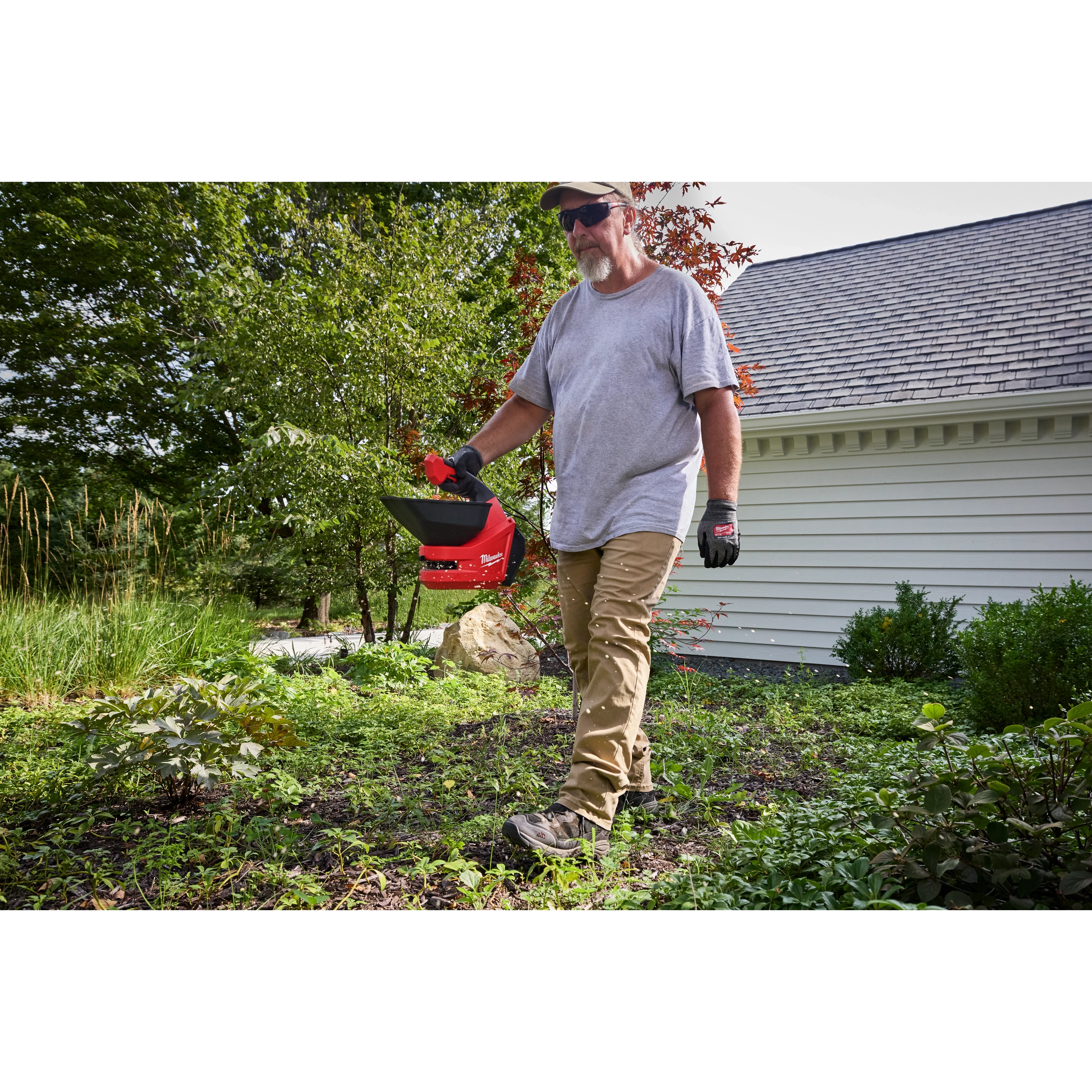 Person walking in a yard holding a red M12 Handheld Broadcast Spreader near a house.