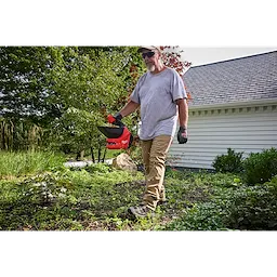 Person walking in a yard holding a red M12 Handheld Broadcast Spreader near a house.