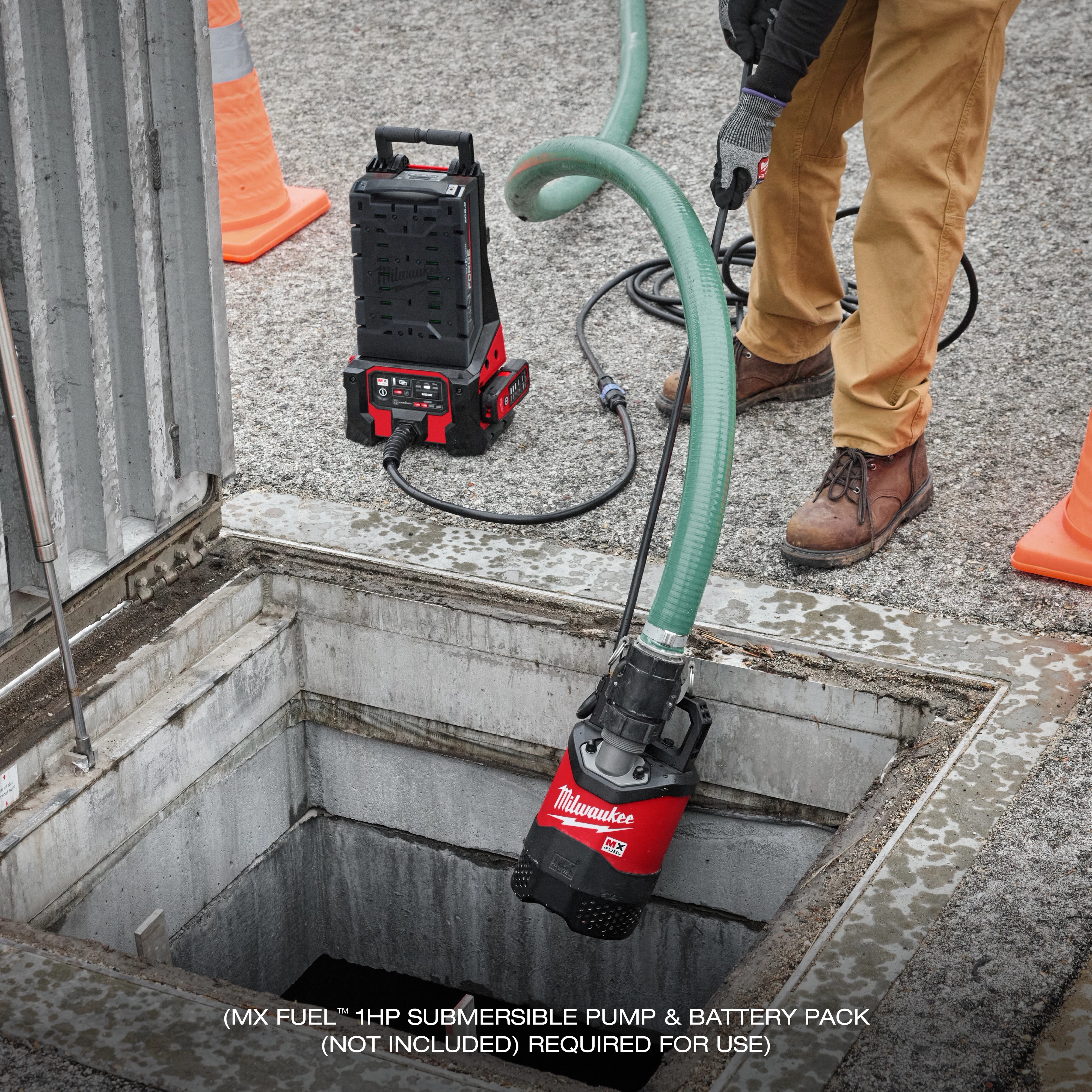 A person operating an MX FUEL™ Portable Pump Power Base, connected to a submersible pump with a green hose, positioned above an open manhole. The power base is placed on the ground nearby. Orange safety cones are in the vicinity.