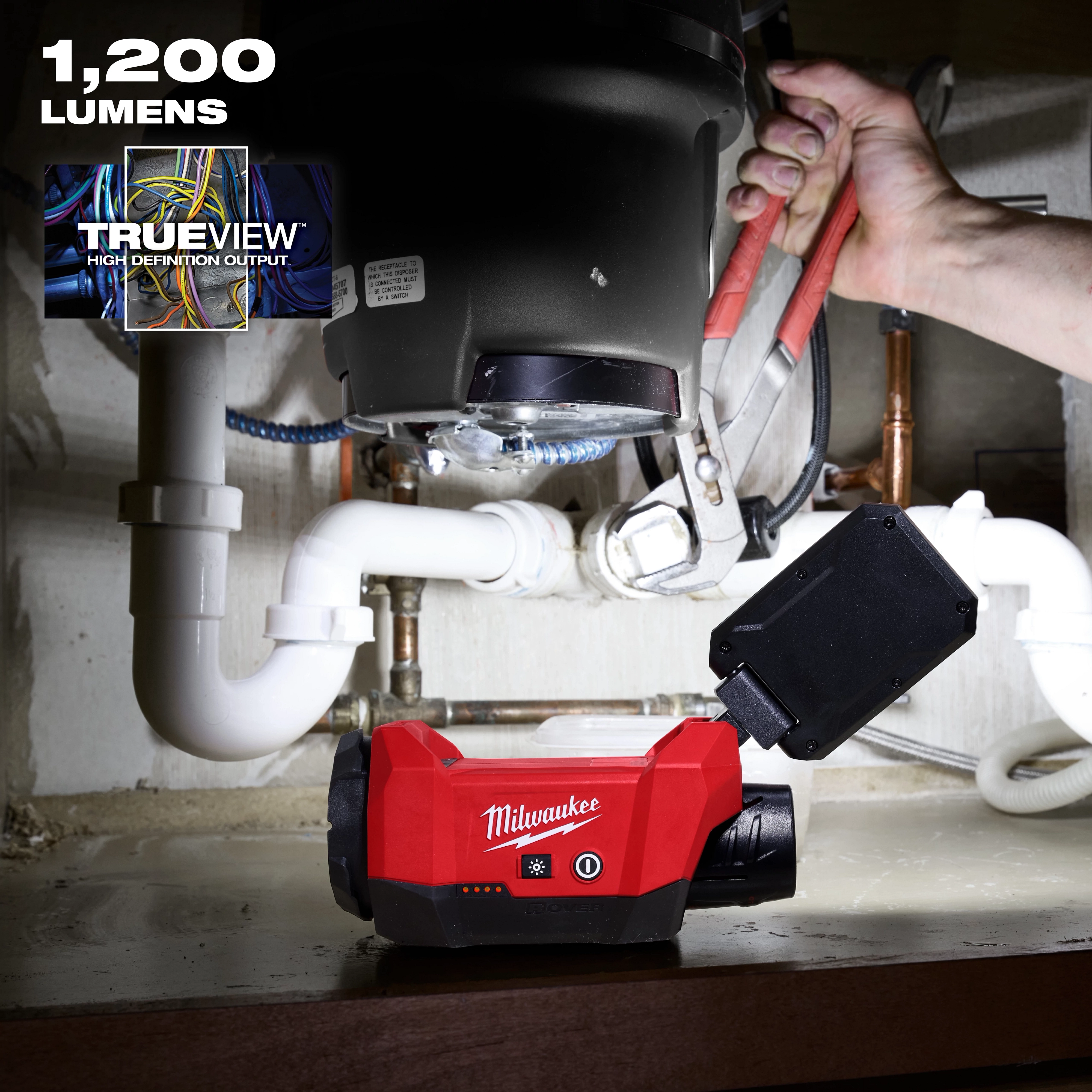 Milwaukee M12™ ROVER™ Multi-Directional Flood Light w/ PACKOUT™ Compatibility is shown illuminating plumbing work under a sink. The red light features TrueView high definition output and delivers 1,200 lumens. A hand is using pliers to work on pipes, benefiting from the versatile light direction.
