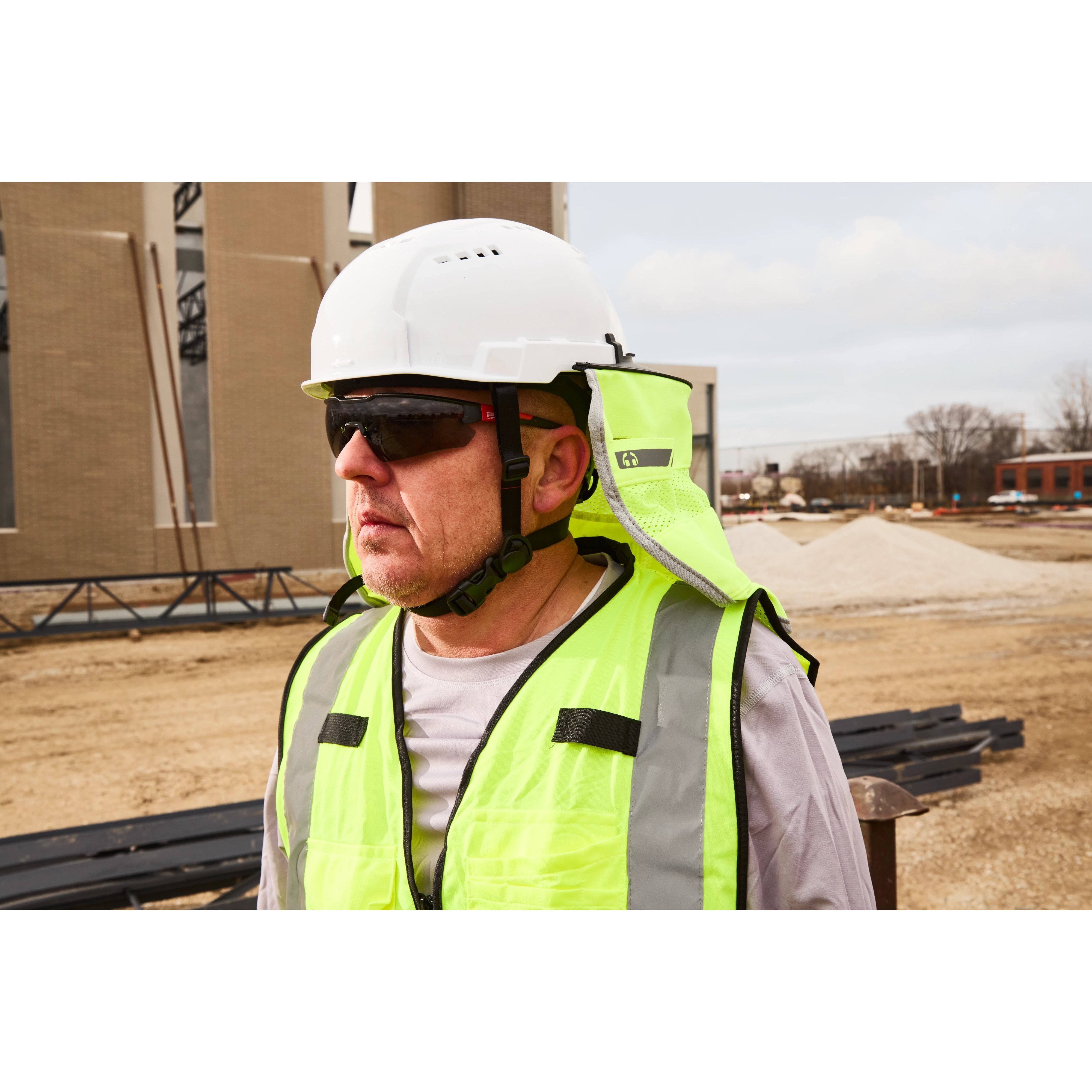 A person is wearing a white hard hat and a high-visibility safety vest at a construction site. The background features a partially constructed building, a pile of gravel, and some bare trees.