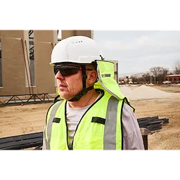 A person is wearing a white hard hat and a high-visibility safety vest at a construction site. The background features a partially constructed building, a pile of gravel, and some bare trees.