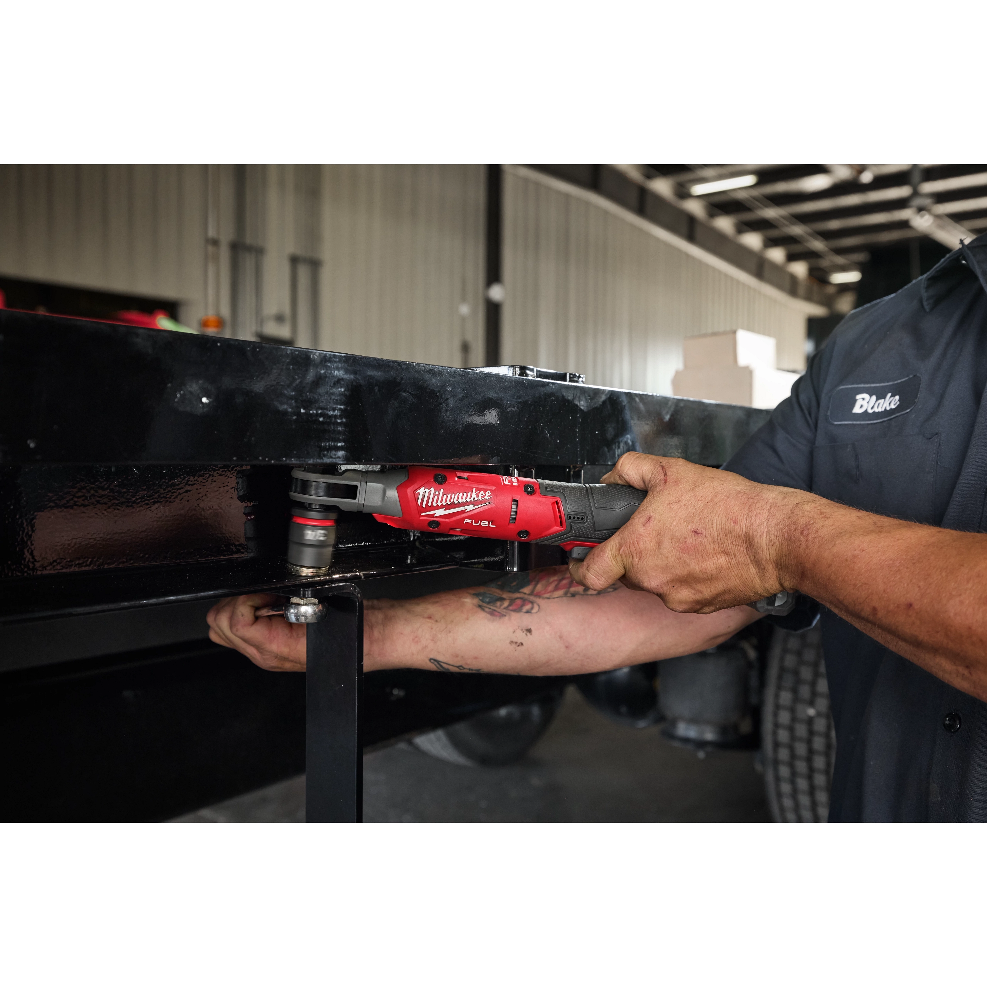 A man named Blake uses the Milwaukee M12 FUEL™ 1/2" Ratchet Kit to tighten a bolt on a metal structure in an industrial setting. His tattooed arm and work shirt are visible, highlighting the tool's compact design and efficient performance in tight spaces.