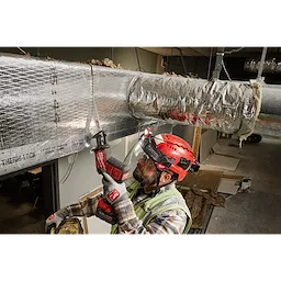 A construction worker wearing a red helmet and safety vest is using a power tool on HVAC ductwork. He is protected by the BOLT™ Gasketed Full Face Shield - Clear Dual Coat Lens, which is compatible with Milwaukee® BOLT™ Safety Helmets & BOLT™ Hard Hats.