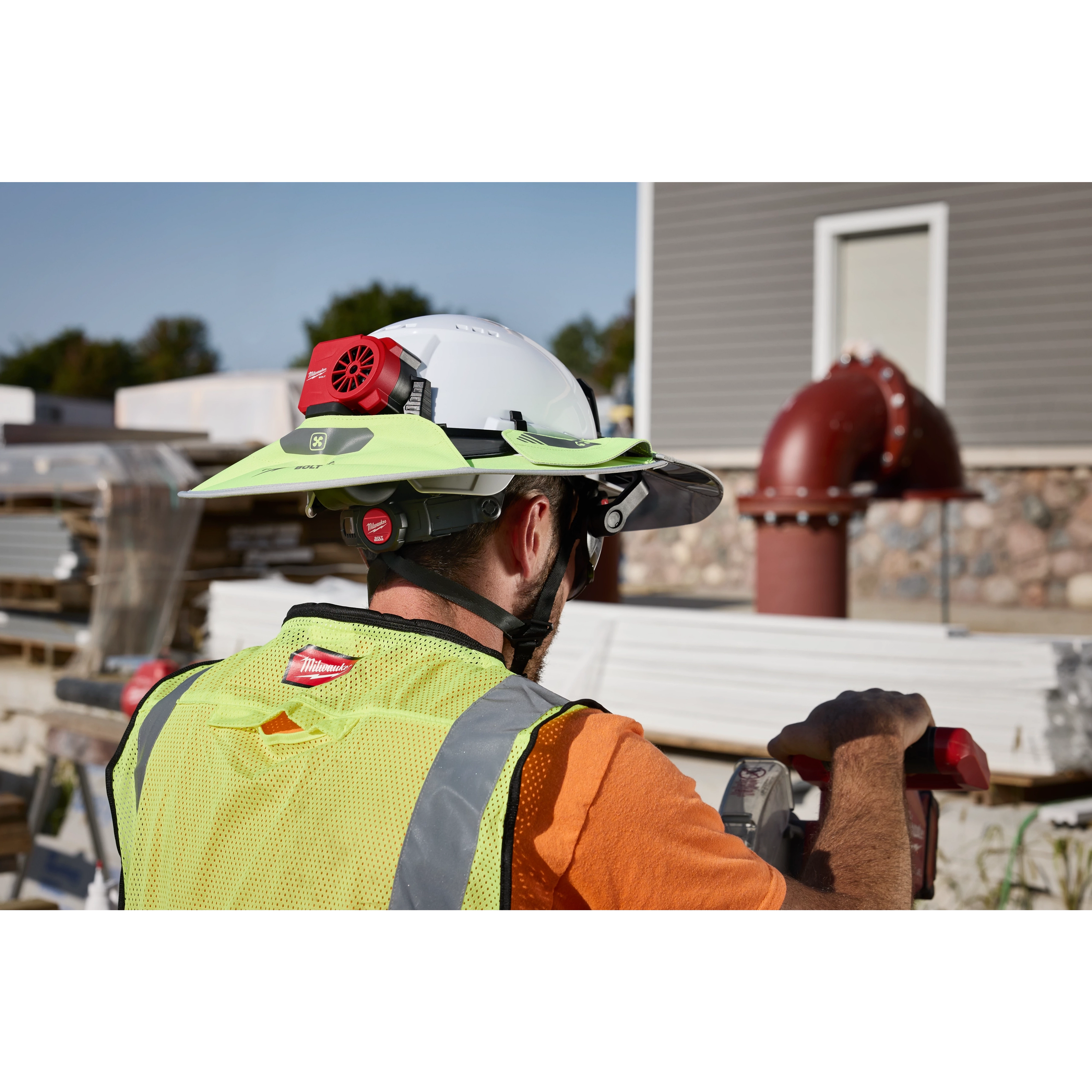A construction worker wearing a white hard hat equipped with a BOLT REDLITHIUM USB Cooling Fan. The background shows a construction site with materials and a red pipe.