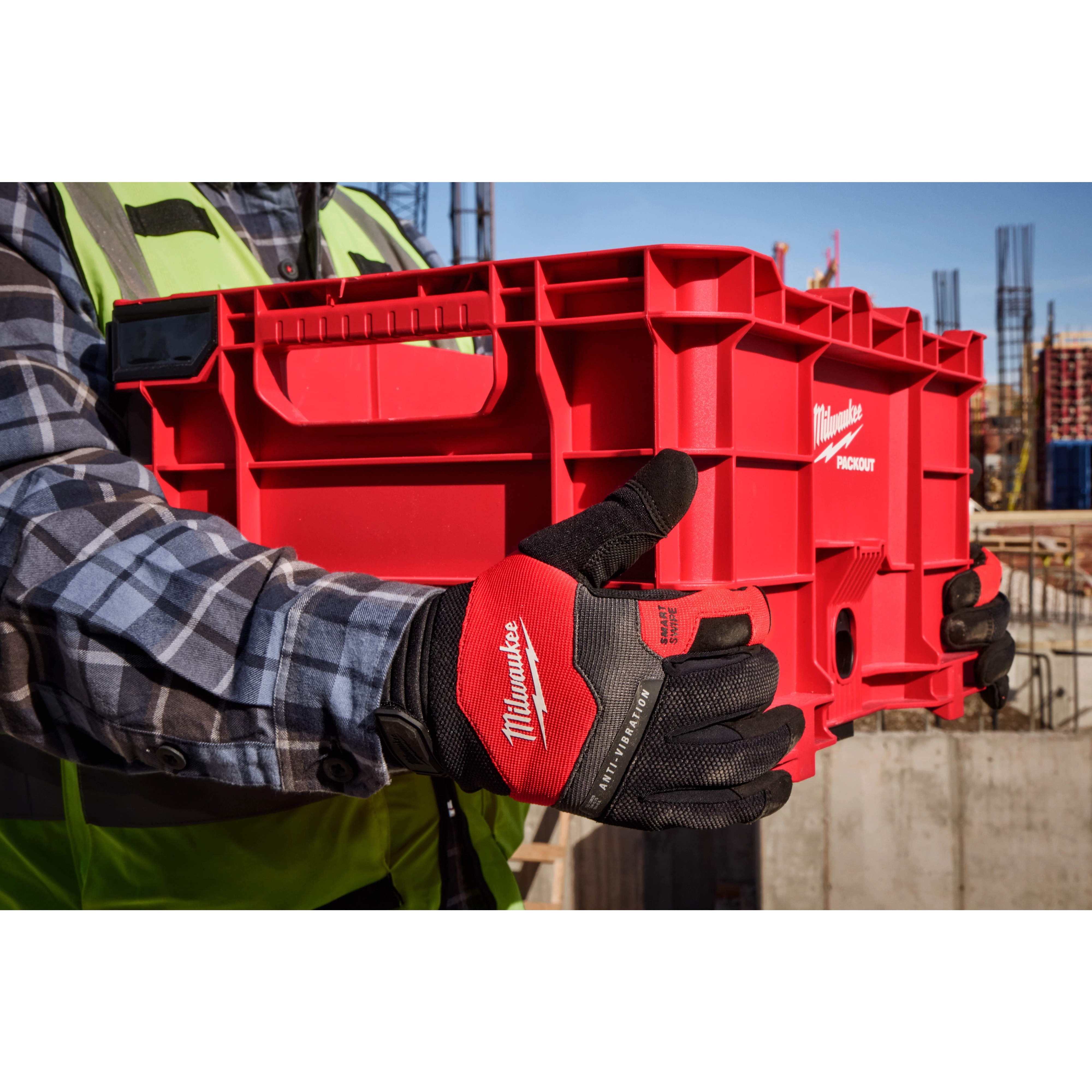 A person wearing Milwaukee-branded Anti-Vibration Work Gloves holds a bright red toolbox. The person is dressed in a plaid shirt and a neon safety vest. The background features a construction site with steel framework and construction materials.