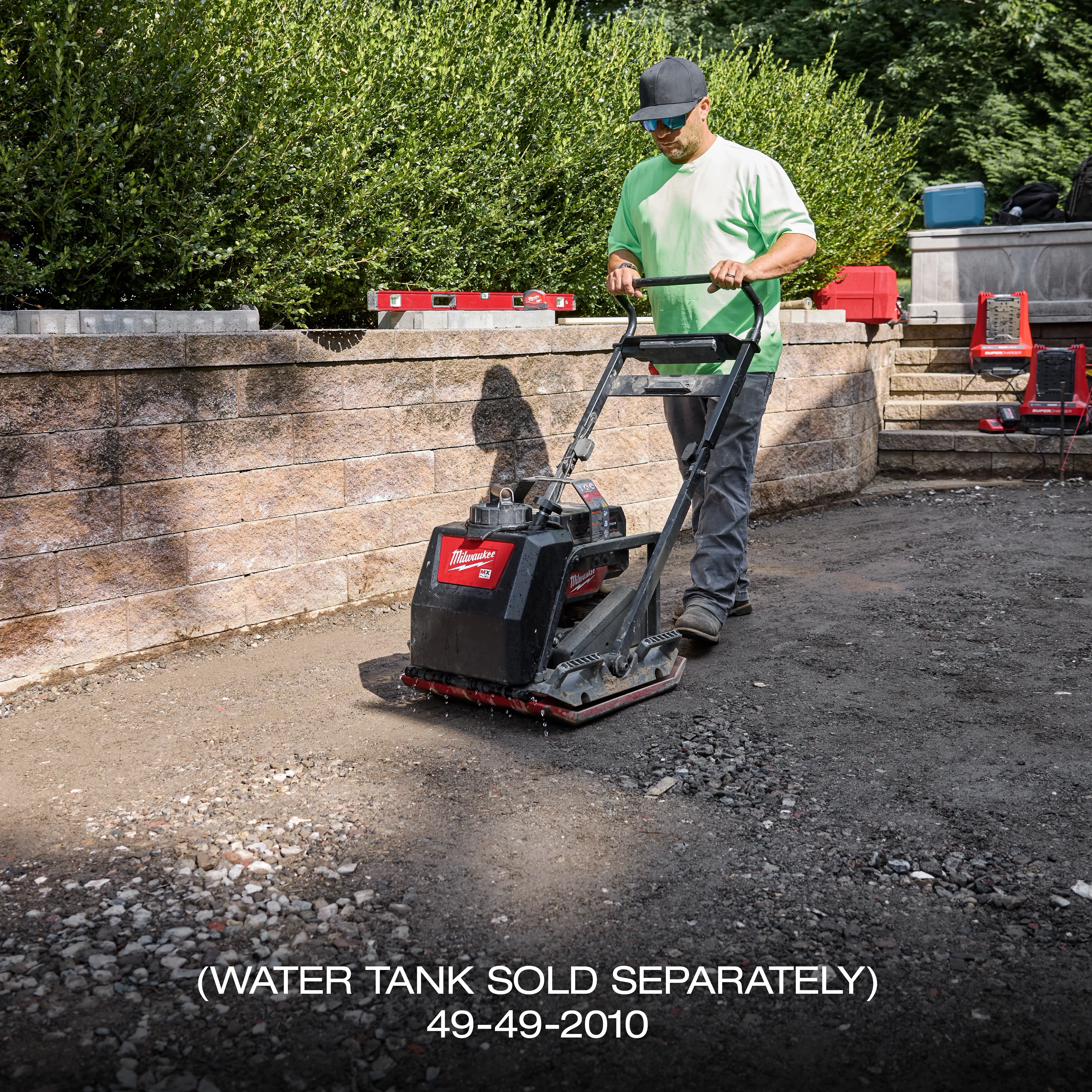 A person uses the MX FUEL™ 20" Plate Compactor Kit on a dirt pathway next to a brick wall and bushes. The compactor has a black and red body with the brand name visible. Various tools and equipment are also visible in the background.