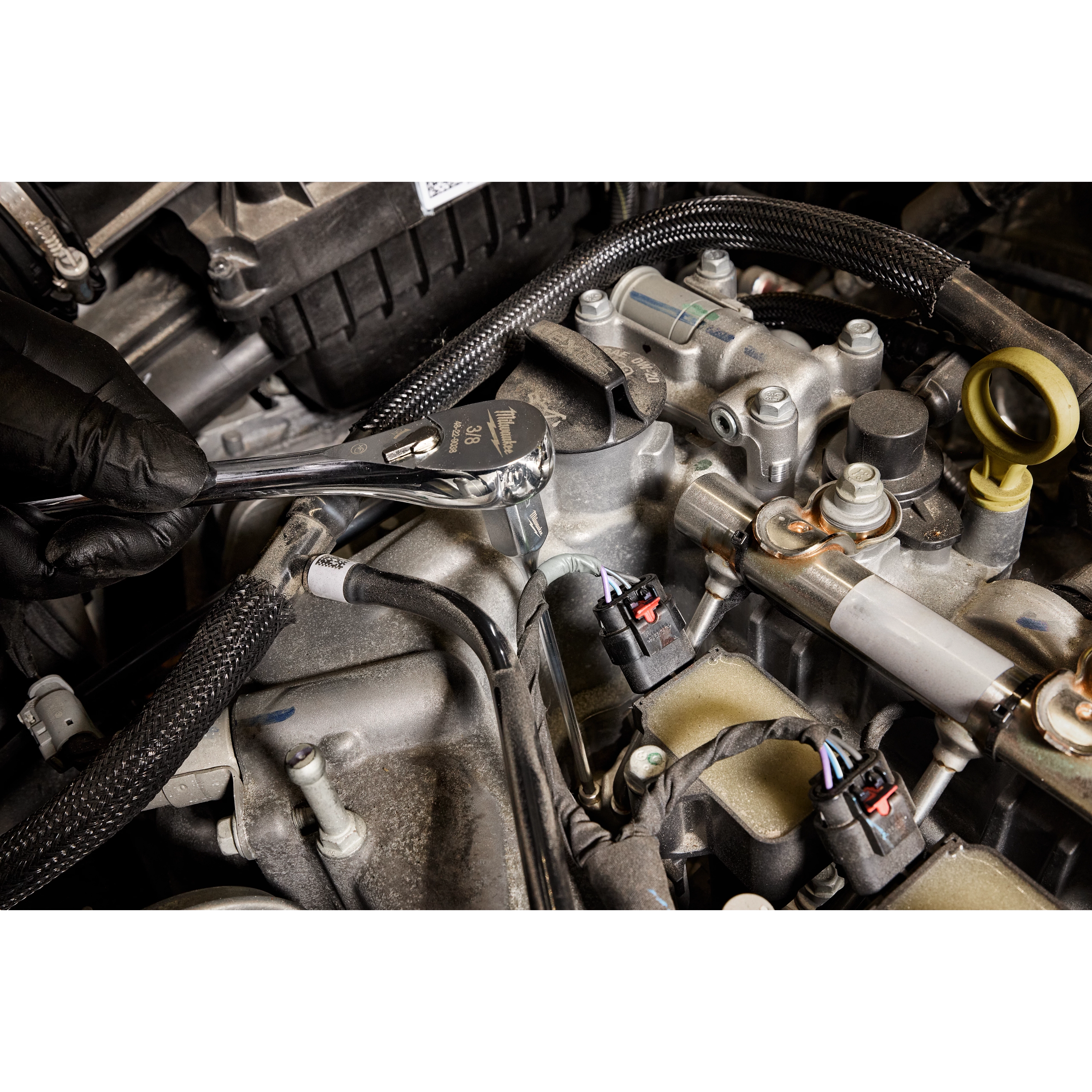 A person wearing a black glove uses a 3/8" Drive 4mm Long Hex Bit Socket attached to a ratchet wrench to work on an engine. The tool is being used to reach a bolt in a tight space within the car's engine compartment.