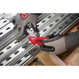 The Multi-function Compact Utility Knife, red with a retractable blade, is held by a gloved hand. Another gloved hand holds a white strip against a metal sheet. The knife is used for precise cutting in construction or DIY projects.