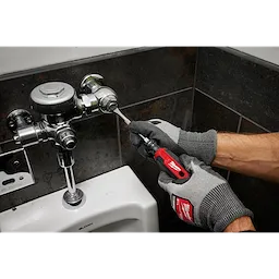 A person wearing gloves uses a 5/16" Slotted 6" Cushion Grip Screwdriver (USA) to adjust a plumbing fixture above a urinal.