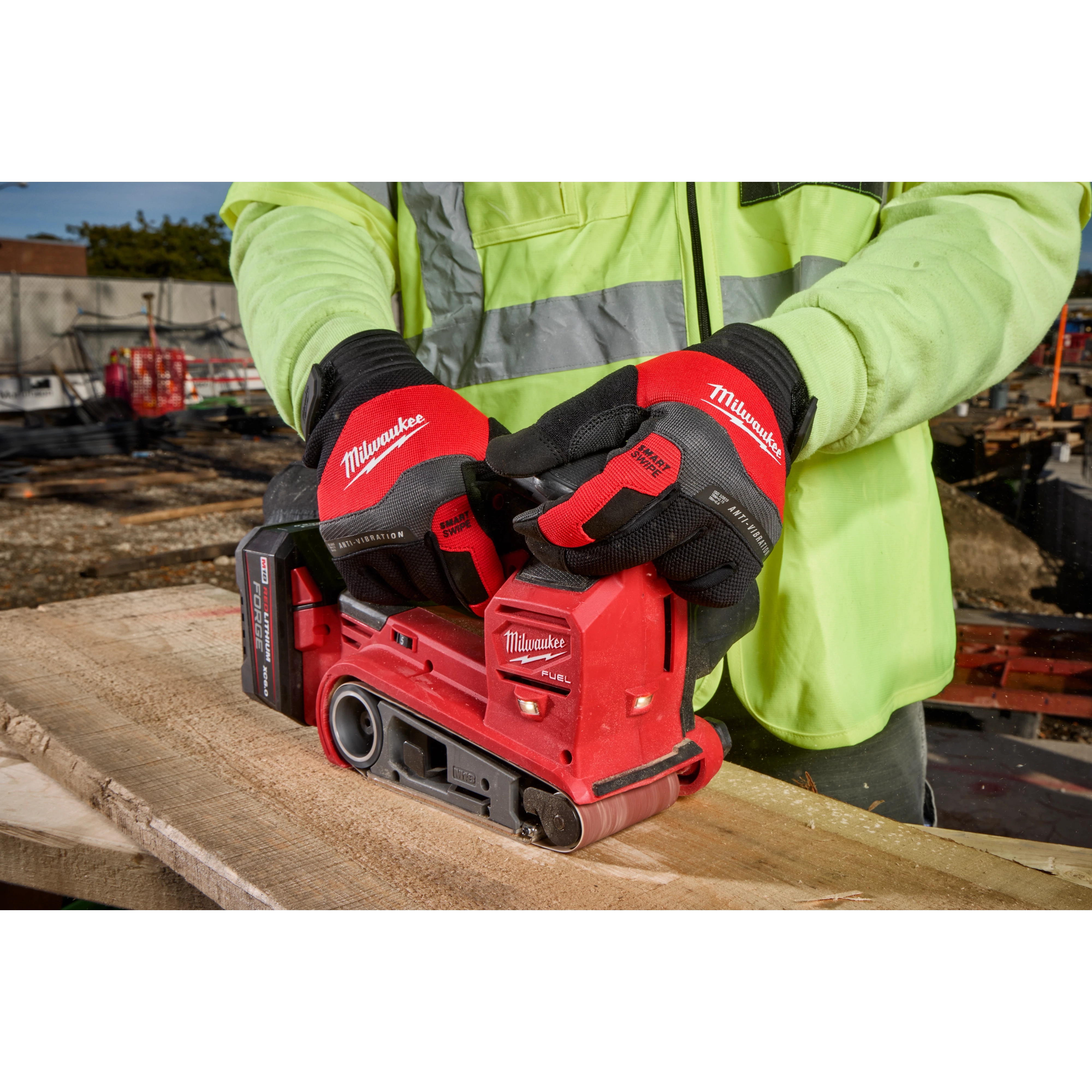 A person wearing Anti-Vibration Work Gloves operates a Milwaukee electric belt sander to smooth a wooden plank at a construction site. The gloves are red and black, designed to minimize vibration and improve grip during tool use. The person is also wearing a high-visibility yellow jacket.