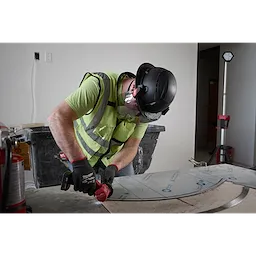 A worker wearing a BOLT™ Black Full Brim Vented Safety Helmet – Type 2, Class C, a high-visibility safety vest, safety gloves, and protective eyewear uses a power tool to work on a metal surface in a construction environment.