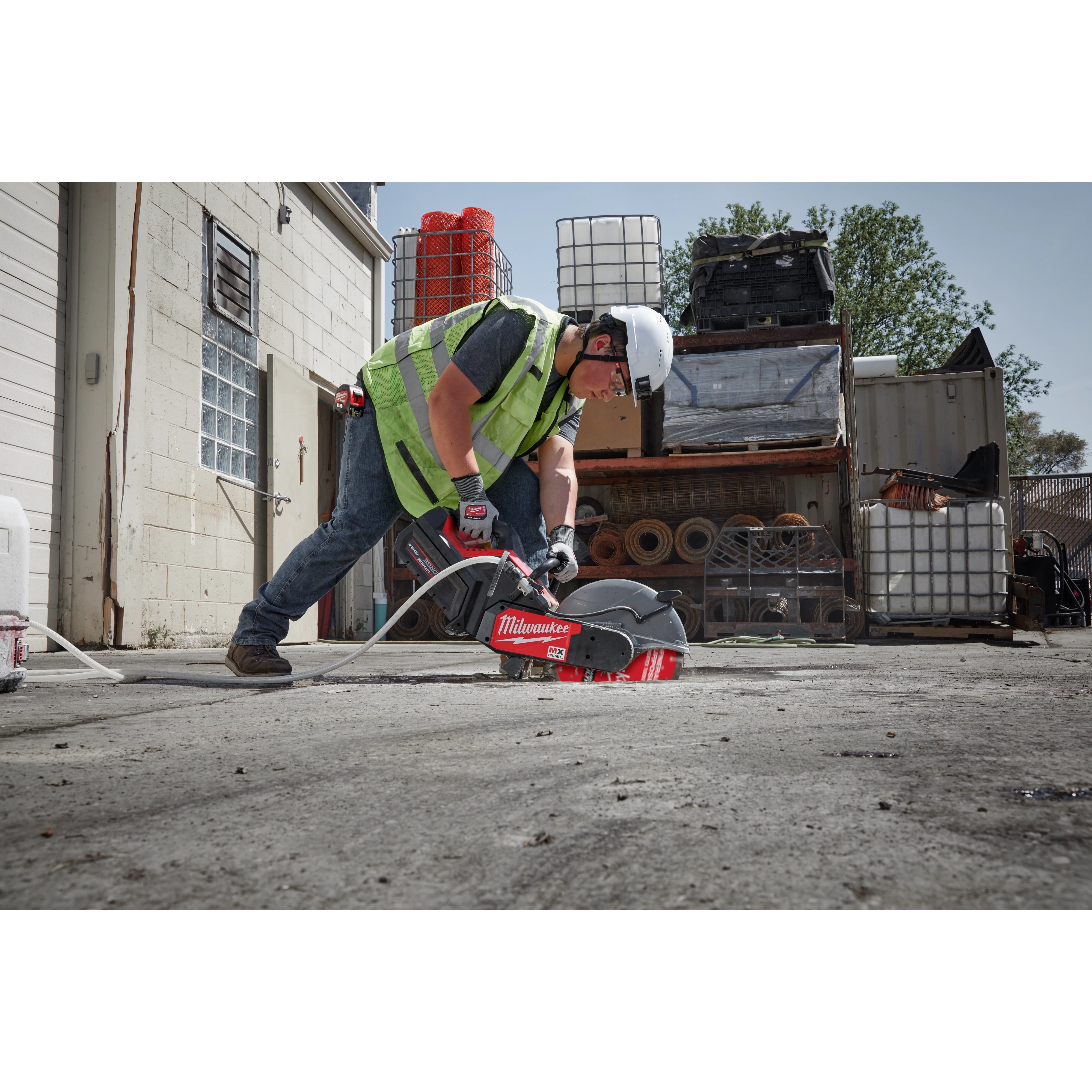 A construction worker using a power saw with a 14" DIAMOND ULTRA™ Segmented Turbo, General Purpose Diamond Blade cuts through concrete in an industrial yard. The worker wears a safety vest, helmet, and protective gloves. Various industrial equipment and materials are stored in the background.