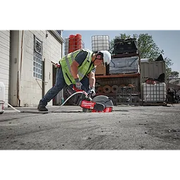 A construction worker using a power saw with a 14" DIAMOND ULTRA™ Segmented Turbo, General Purpose Diamond Blade cuts through concrete in an industrial yard. The worker wears a safety vest, helmet, and protective gloves. Various industrial equipment and materials are stored in the background.