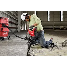 A worker in a construction site uses the M18™ FUEL™ 1-9/16" SDS Max Rotary Hammer w/ ONE-KEY™ to break up concrete. He wears a white hard hat, ear protection, gloves, and safety glasses. The device is connected to a dust extraction system to maintain cleanliness.