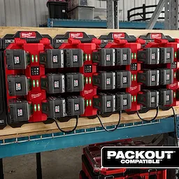 The image shows an M18™ Six Bay Daisy Chain Rapid Charger mounted on a wooden surface. The charger is holding multiple battery packs with green indicator lights illuminated. The charger setup is labeled "PACKOUT™ COMPATIBLE" at the bottom right of the image.