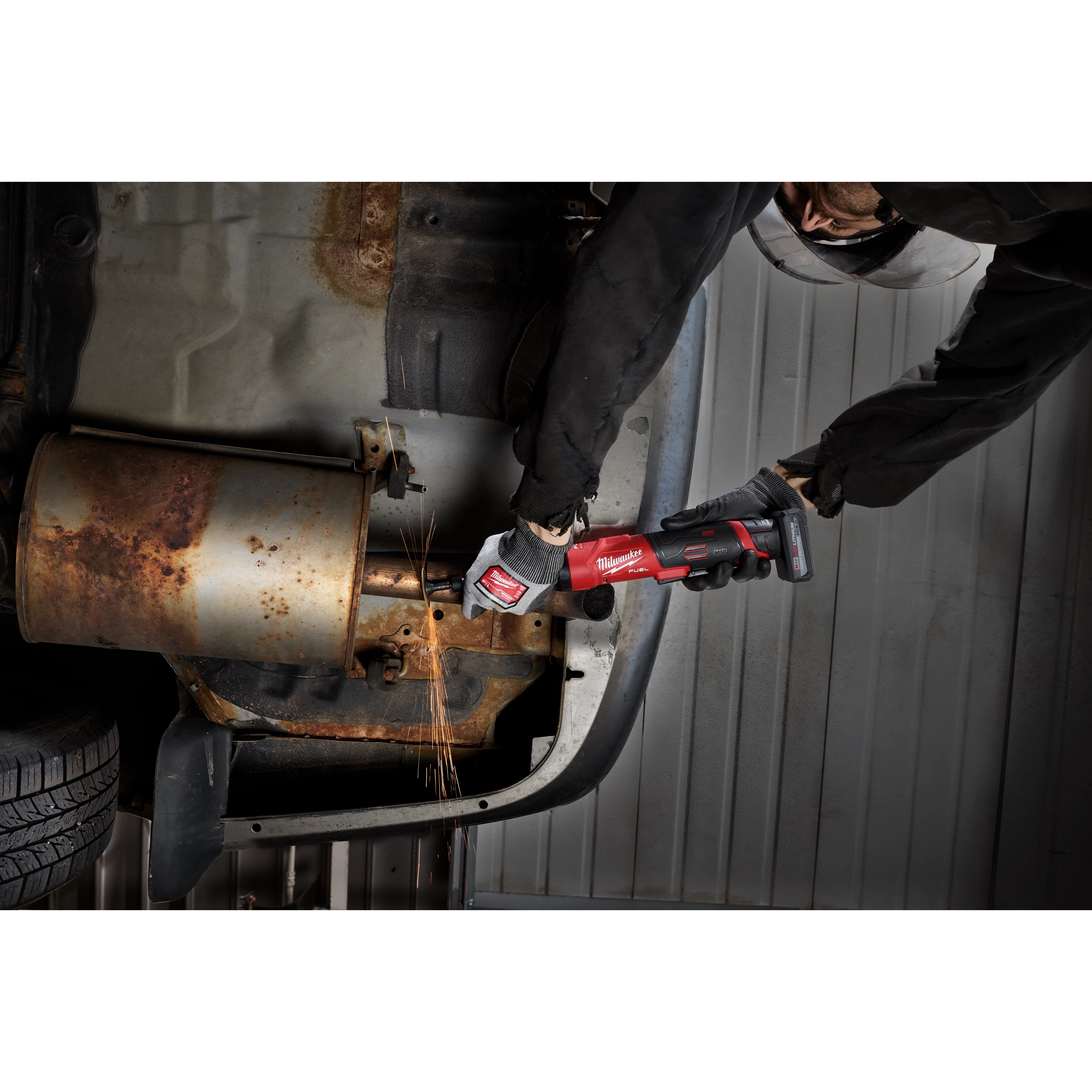 Worker using an M12 FUEL 1/4" Extended Die Grinder on rusted metal under a vehicle.