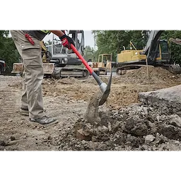 A person uses a 5lb Pick Mattock with a 36" handle to break up hard ground at a construction site. Heavy machinery and earth movers are visible in the background. The user wears protective gloves and work pants.