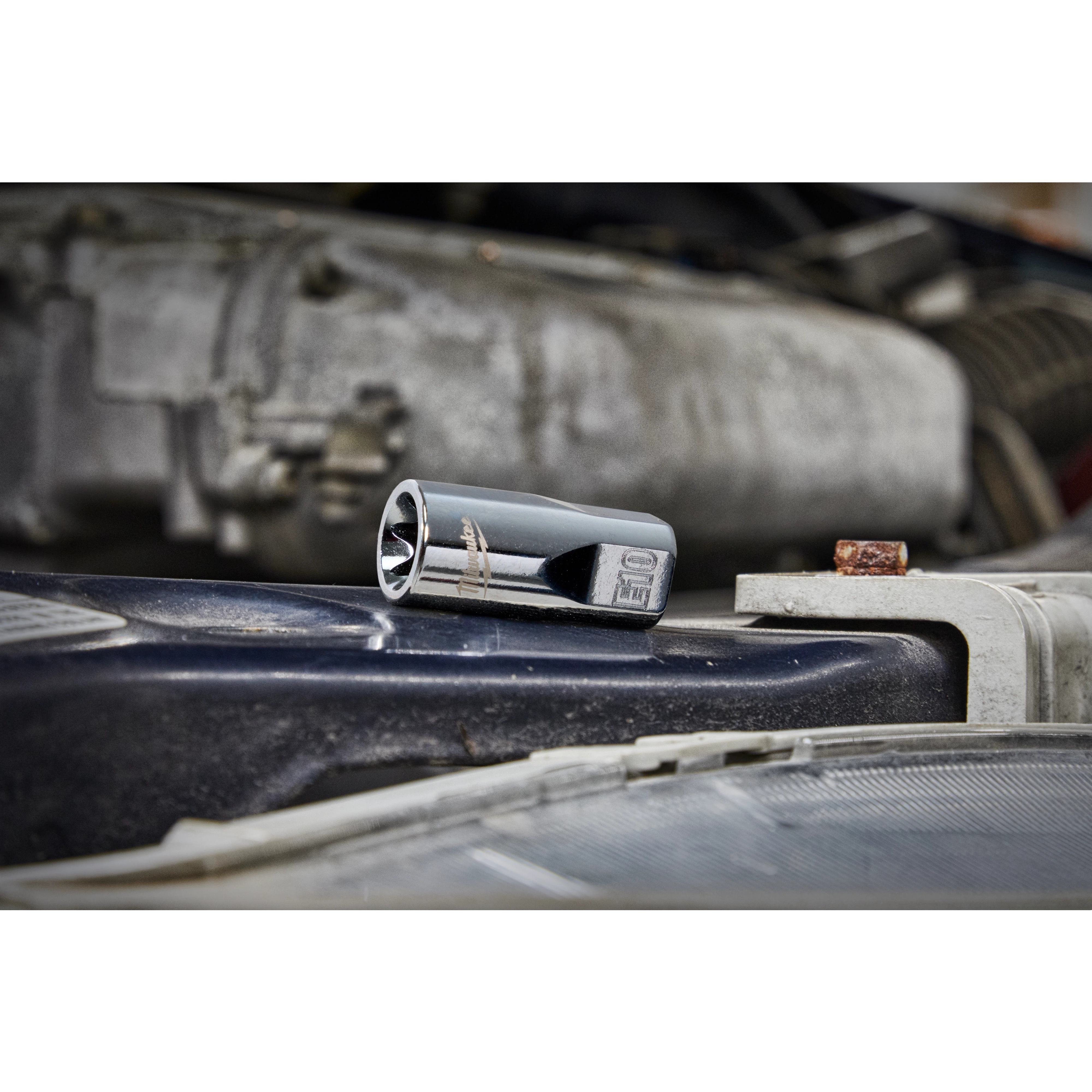 A shiny E10 socket rests on a dusty car engine part. The background includes bright, blurry components of the engine. The image focuses on the socket tool, illustrating its reflective chrome surface with other elements slightly out of focus.