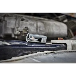 A shiny E10 socket rests on a dusty car engine part. The background includes bright, blurry components of the engine. The image focuses on the socket tool, illustrating its reflective chrome surface with other elements slightly out of focus.