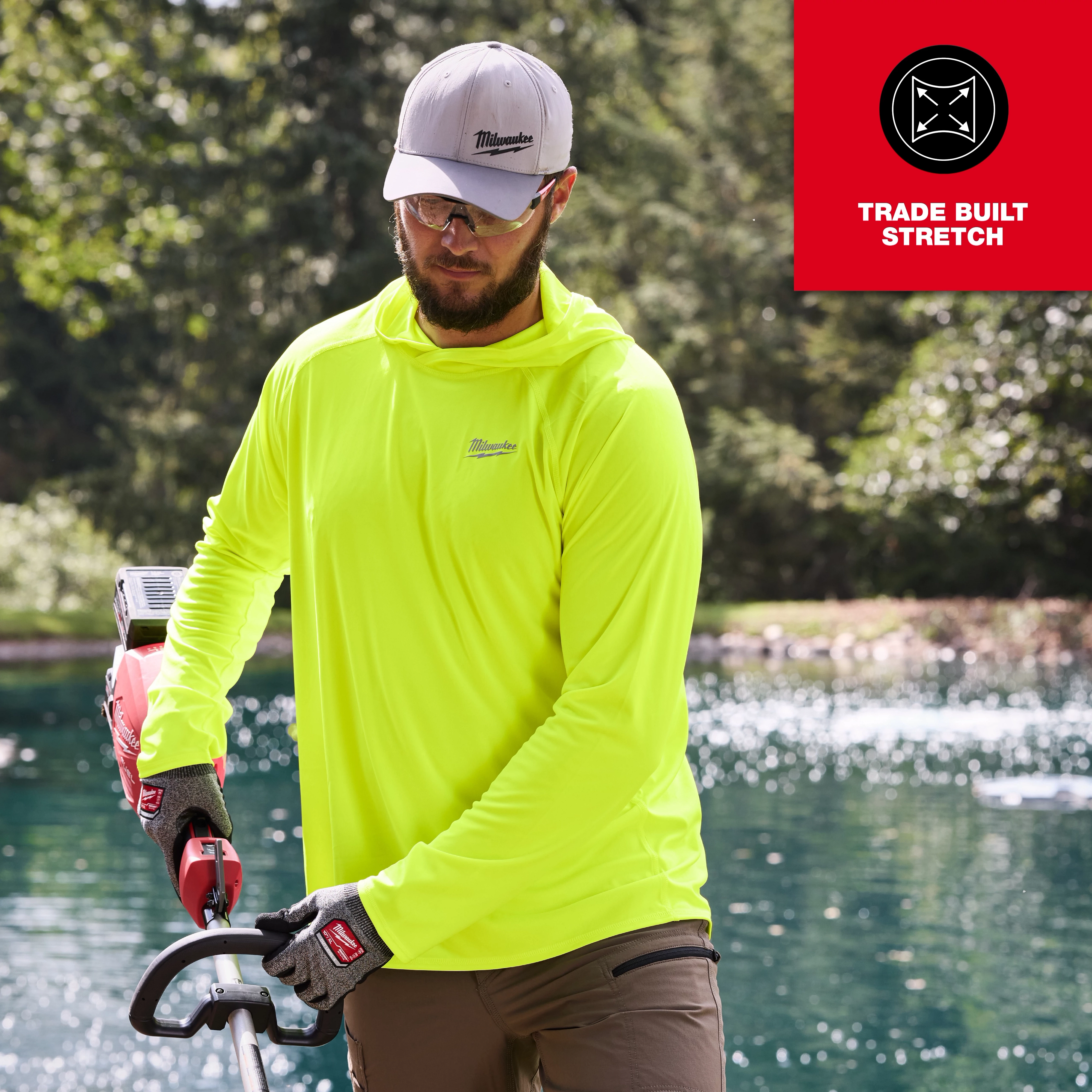 A person wearing a bright yellow WORKSKIN Hooded Sun Shirt and gloves is using a power tool near a pond. A "Trade Built Stretch" emblem is displayed in the top right corner.