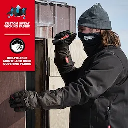 Person in cold weather gear works with a flashlight by a wooden structure. Wearing WORKSKIN™ Balaclava beneath a beanie. The product is highlighted for its custom sweat-wicking and breathable mouth and nose covering fabric.