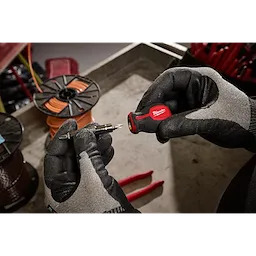 A person wearing gloves is holding a 6-IN-1 Stubby Multi-Bit Screwdriver in one hand and a detachable bit in the other. The background includes tools and wire spools.