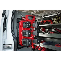 The image shows an M18™ Six Bay Daisy Chain Rapid Charger mounted inside a vehicle. The charger, red in color, is holding six batteries and is secured to the wall. Shelving units and various tools are visible in the background inside the vehicle.