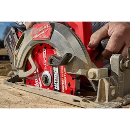 A Milwaukee Fuel circular saw is shown cutting wood, featuring the 7-1/4” 24T NITRUS™ Carbide Framing & Demolition Circular Saw Blade. A worker's hand is positioned on the saw's handle as it actively cuts through the material. The blade is red with branding and specifications visible.