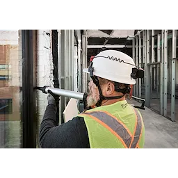 A construction worker wearing a BOLT 4PT White Front Brim Non-Vented Safety Helmet, a hi-vis vest, and using a power tool on a building frame.