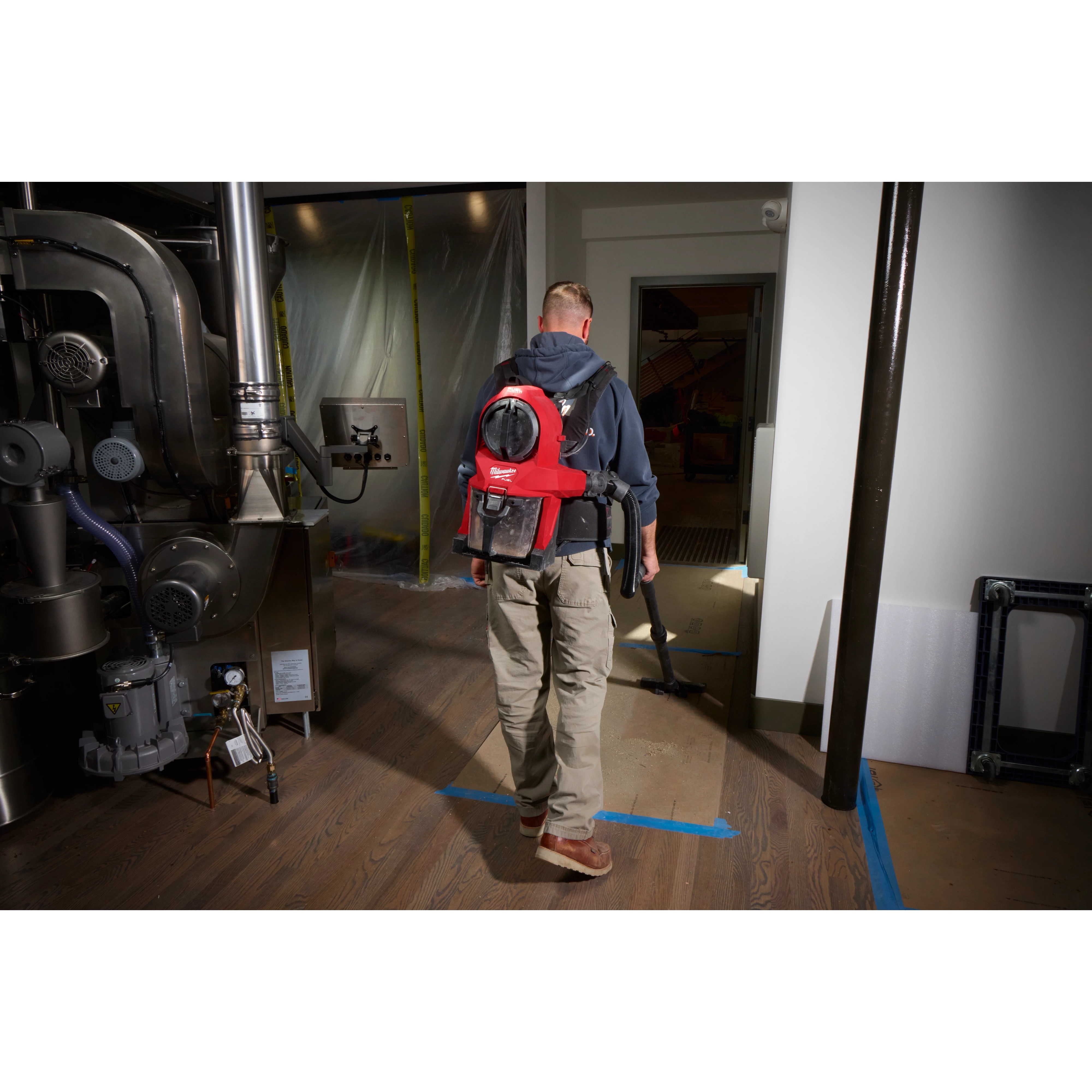 Man using the M18 FUEL™ 3-in-1 Backpack Vacuum to clean a hardwood floor in an industrial setting. The vacuum is red and black, strapped to his back, and connected through a long hose. The room contains large machinery and is partially under construction.
