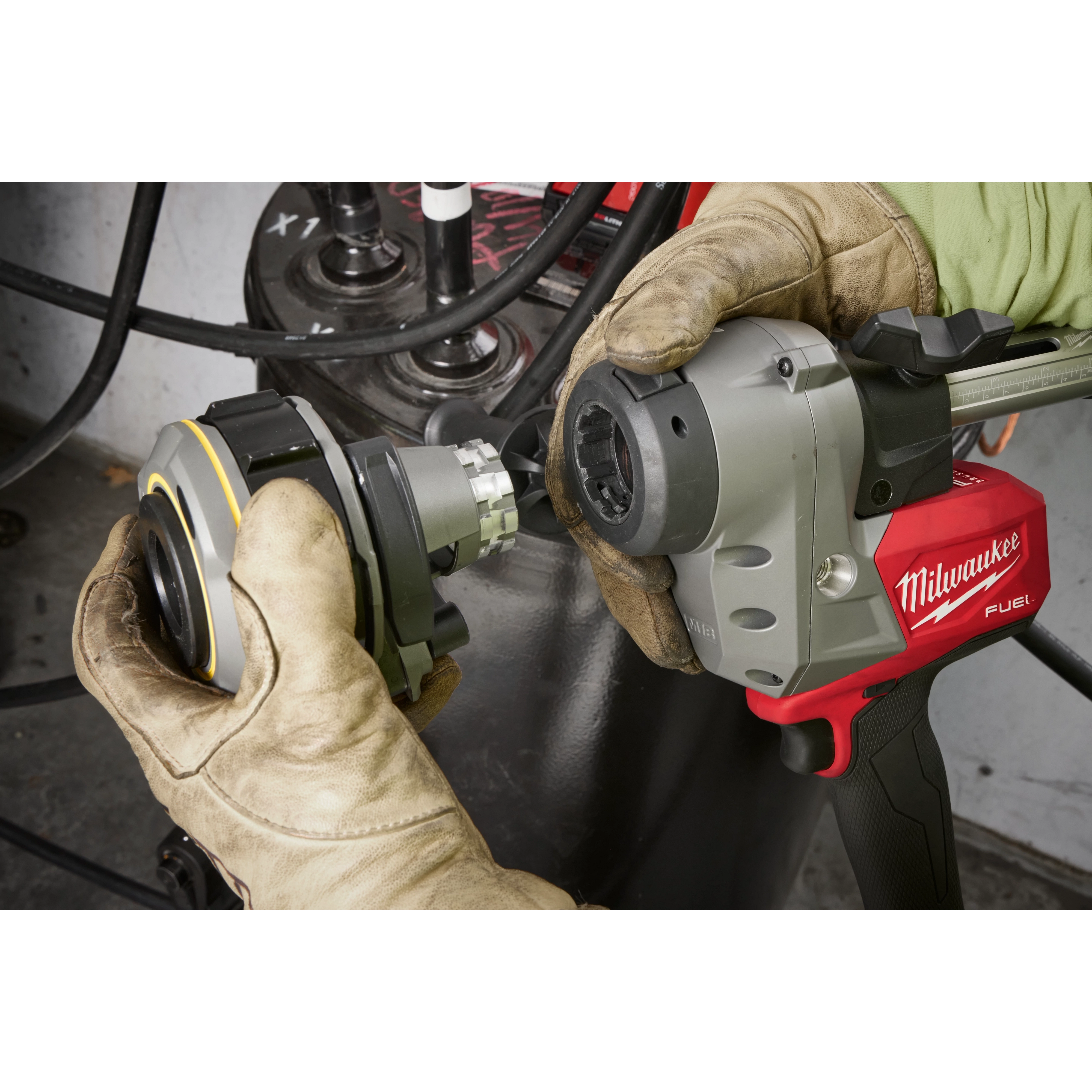 The M18™ FUEL™ Covered Conductor Cable Stripper by Milwaukee is held by gloved hands during operation. The red and gray power tool appears to be in use with a metallic fitting attachment being connected. The background features cables and industrial equipment.
