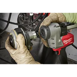 The M18™ FUEL™ Covered Conductor Cable Stripper by Milwaukee is held by gloved hands during operation. The red and gray power tool appears to be in use with a metallic fitting attachment being connected. The background features cables and industrial equipment.