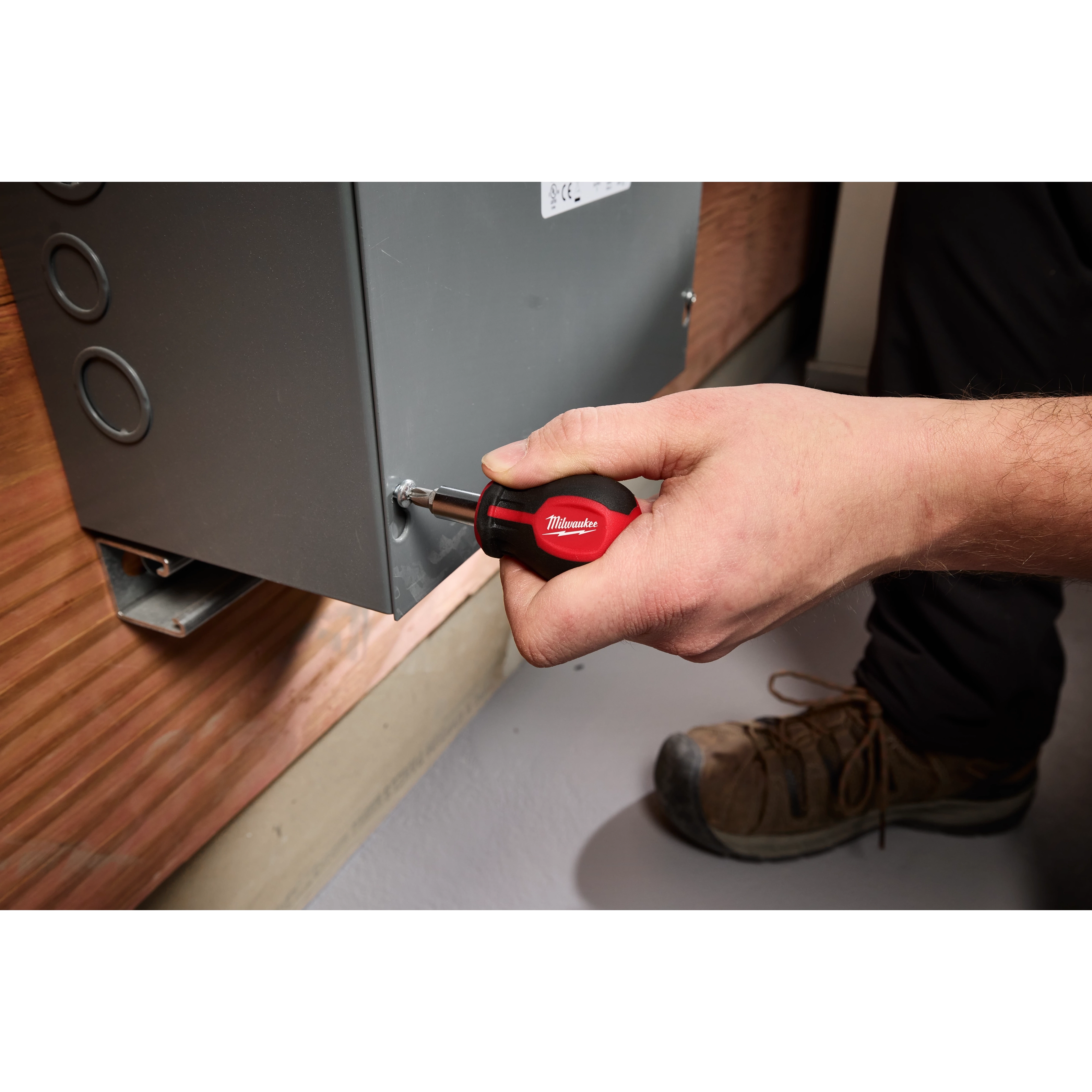 A person uses a 6-IN-1 Stubby Multi-Bit Screwdriver to tighten a screw on a metal electrical panel. The screwdriver has a red and black handle, with visible branding.