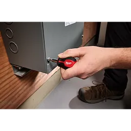 A person uses a 6-IN-1 Stubby Multi-Bit Screwdriver to tighten a screw on a metal electrical panel. The screwdriver has a red and black handle, with visible branding.