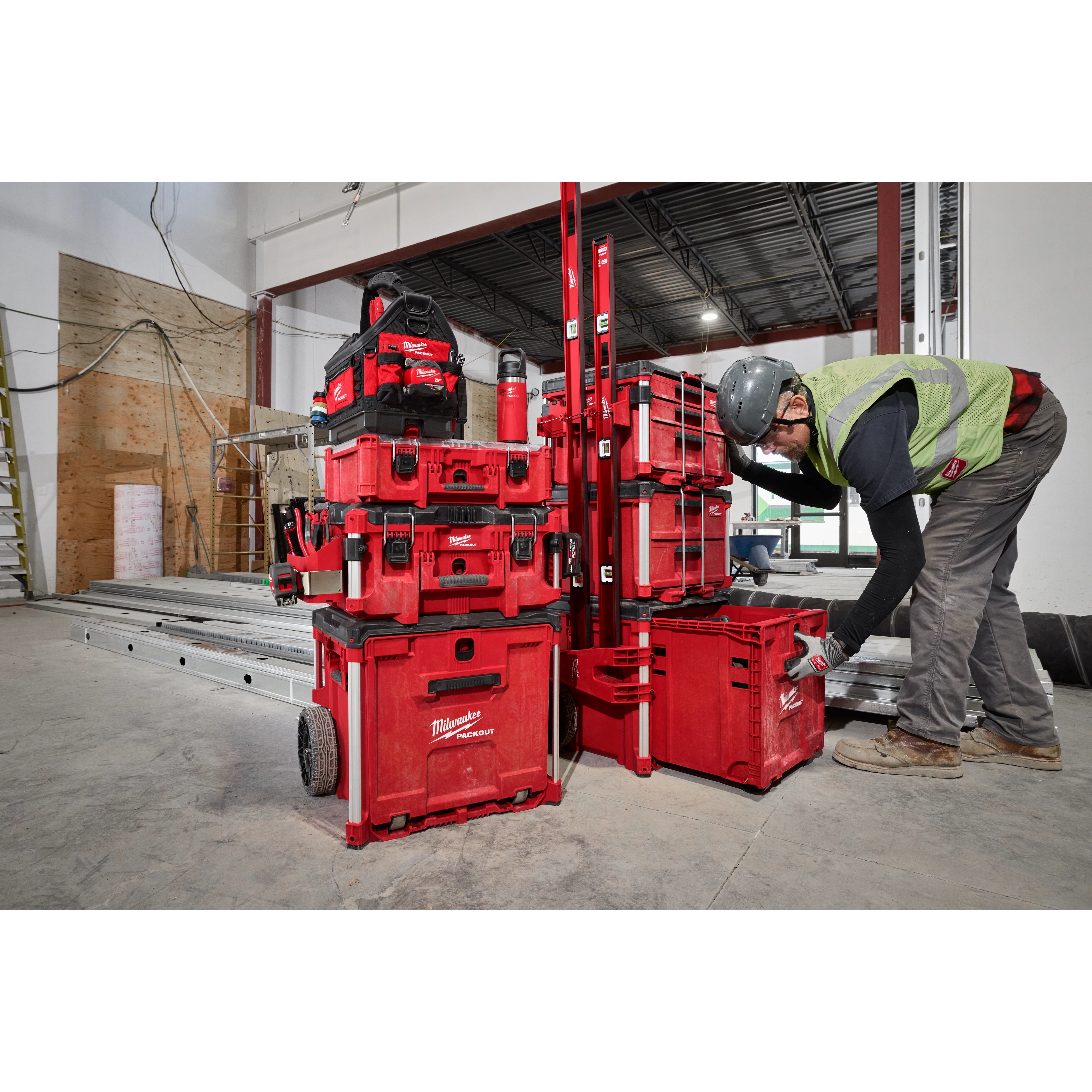 A construction worker in safety gear organizes tools using a stack of red PACKOUT™ Rolling Drawer Tool Boxes in a partially built interior space with equipment and materials nearby.