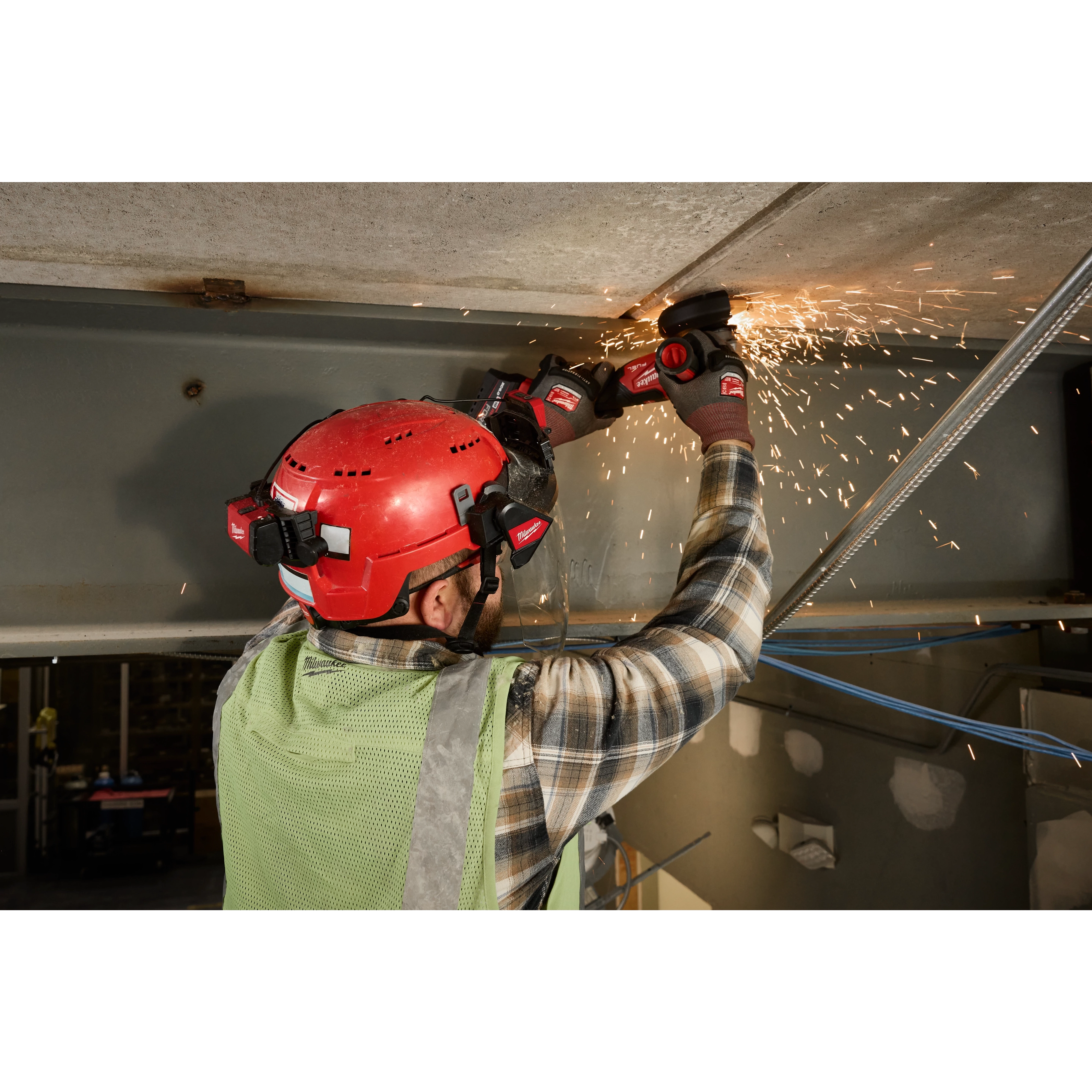 A worker wearing a red Milwaukee® safety helmet with the BOLT™ Gasketed Full Face Shield - Clear Dual Coat Lens and a green vest grinds a metal surface, producing sparks. The face shield is compatible with Milwaukee® BOLT™ Safety Helmets & BOLT™ Hard Hats.