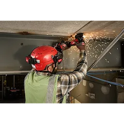 A worker wearing a red Milwaukee® safety helmet with the BOLT™ Gasketed Full Face Shield - Clear Dual Coat Lens and a green vest grinds a metal surface, producing sparks. The face shield is compatible with Milwaukee® BOLT™ Safety Helmets & BOLT™ Hard Hats.