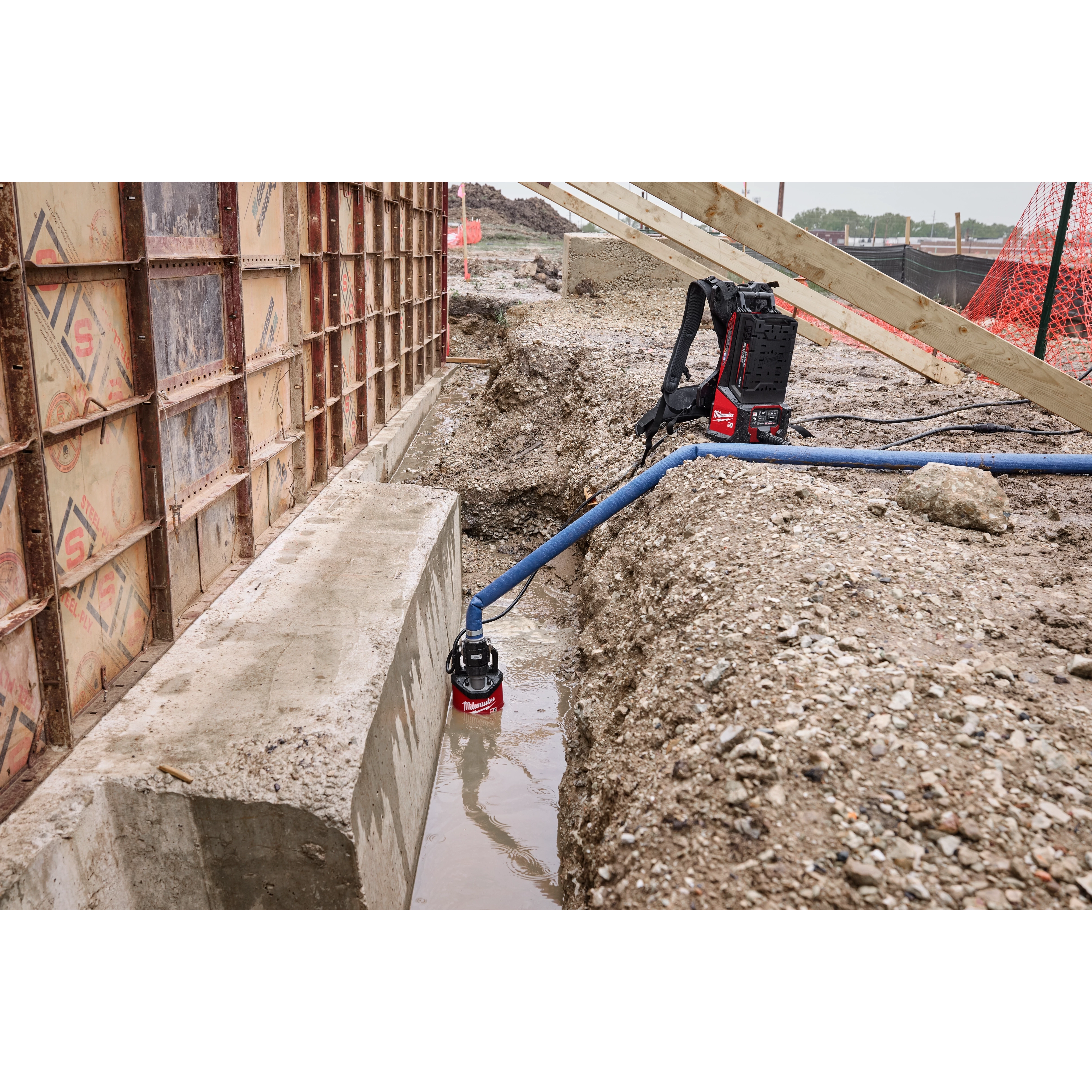 An MX FUEL™ Portable Pump Power Base and an MX FUEL™ 1HP 2" Submersible Pump Kit in use at a construction site. The submersible pump is placed in a water-filled trench, connected to the power base via a blue hose.