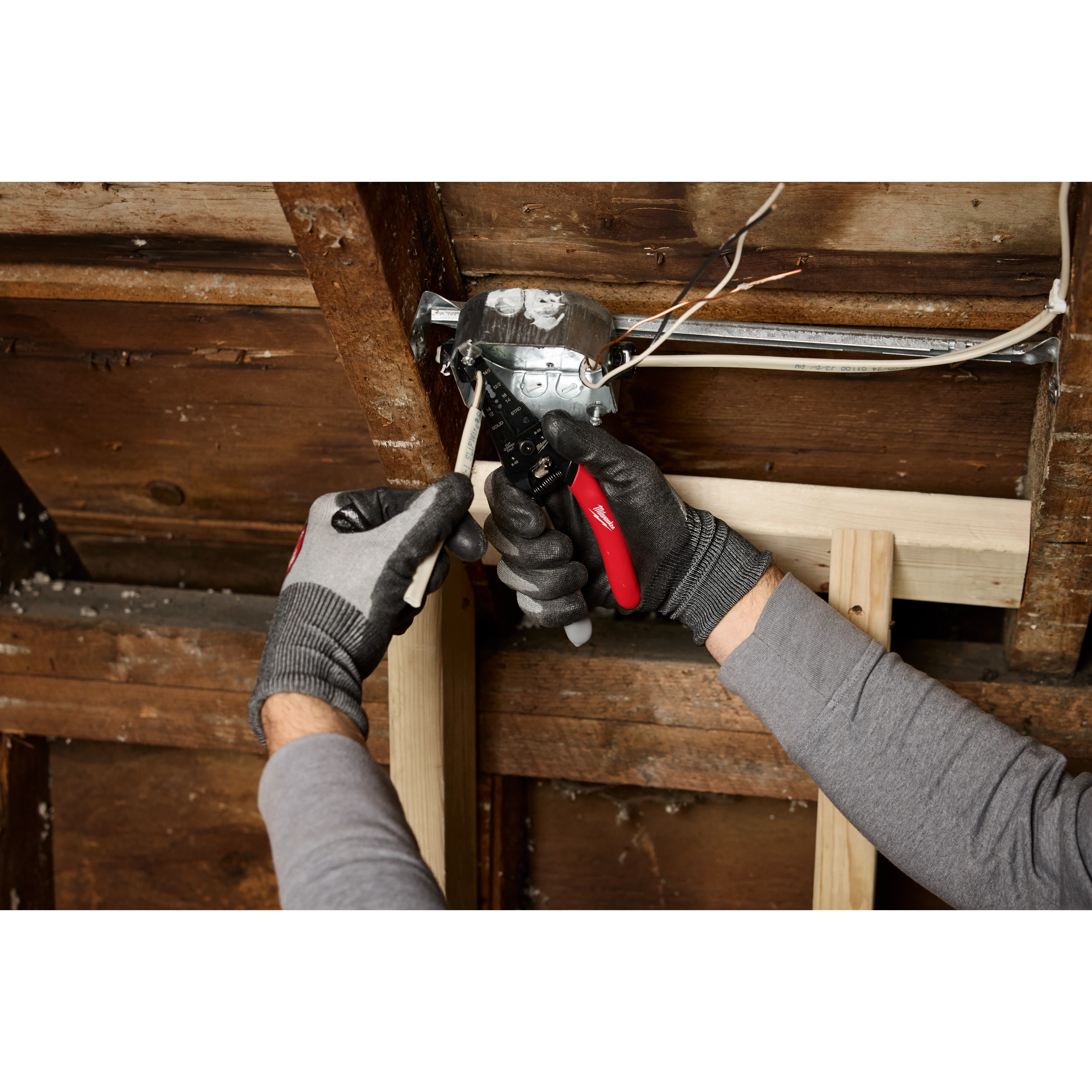 A person wearing gloves is using a 12/2 & 14/2 NM Dipped Grip Wire Stripper & Cutter to strip wires inside a partially installed electrical box in a wooden structure.
