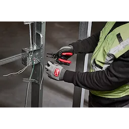 A person wearing gloves and a safety vest uses a 10-24 AWG Compact Dipped Grip Wire Stripper & Cutter to work on electrical wires attached to a metal structure inside an unfinished building.