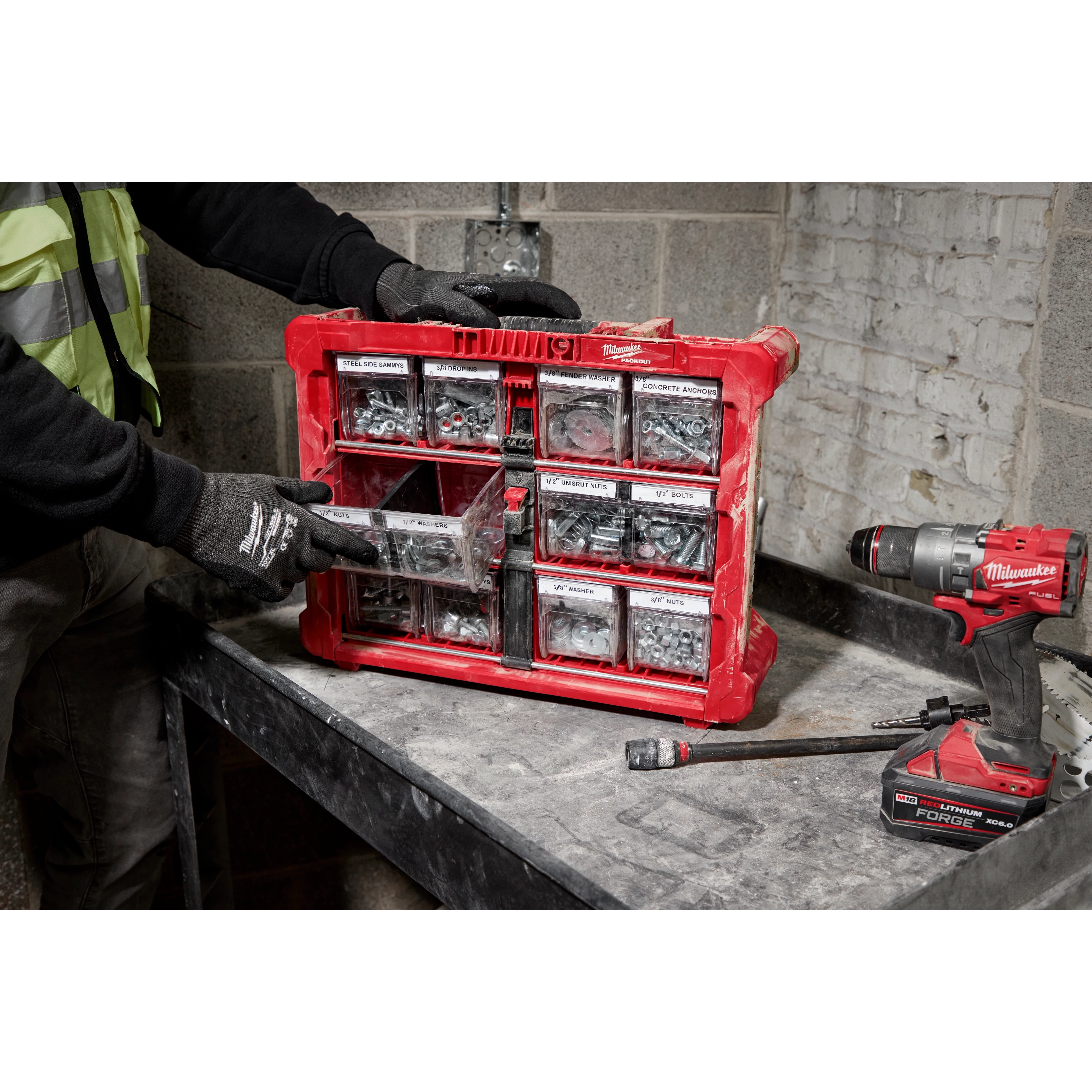 A person wearing gloves organizes a red portable tool organizer box containing various screws and fasteners. The box is placed on a workbench next to a Milwaukee cordless drill and a hammer. The background features a concrete and brick wall.